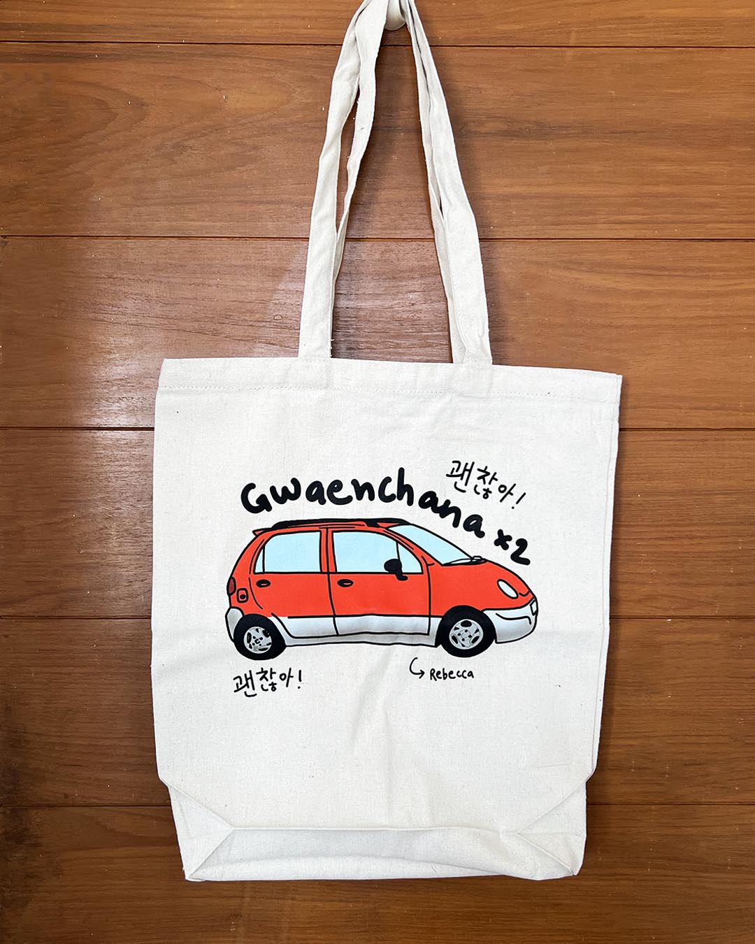 White tote bag with a red car illustration and text 'gwaenchana' and Rebecca on a wooden surface