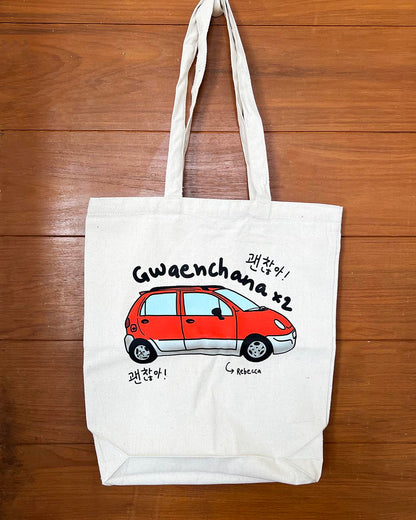 White tote bag with a red car illustration and text 'gwaenchana' and Rebecca on a wooden surface