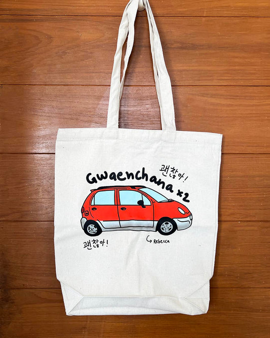 White tote bag with a red car illustration and text 'gwaenchana' and Rebecca on a wooden surface